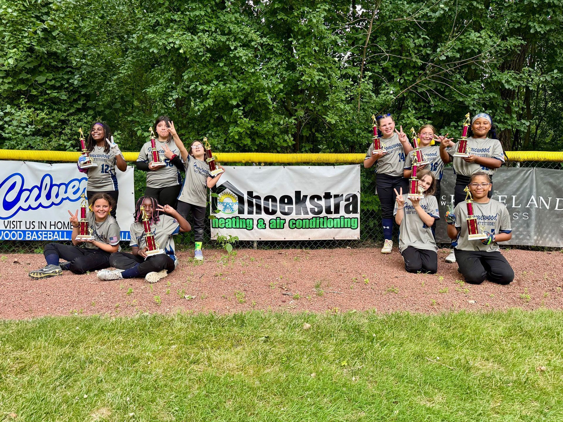 Youth softball team holding trophies, smiling, on a field with sponsor banners in the background.