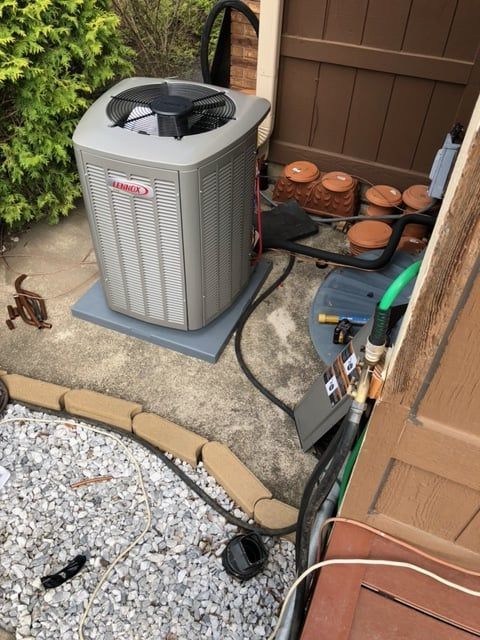 A Lennox air conditioning unit on a concrete pad near a brown fence, with tools and a hose nearby.