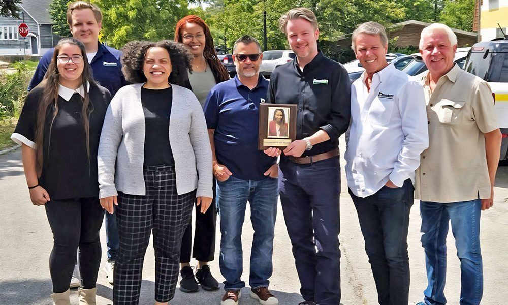 Group of people posing outdoors, some holding a framed photo. Sunny day, parking lot.