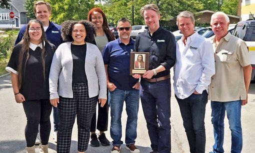 Group of people outdoors holding a framed image. Some smiling, sunny day. Buildings and cars visible.