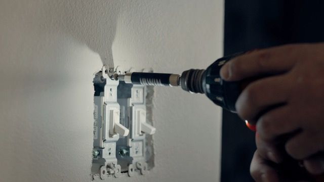 A person is using a drill to install a light switch on a wall.