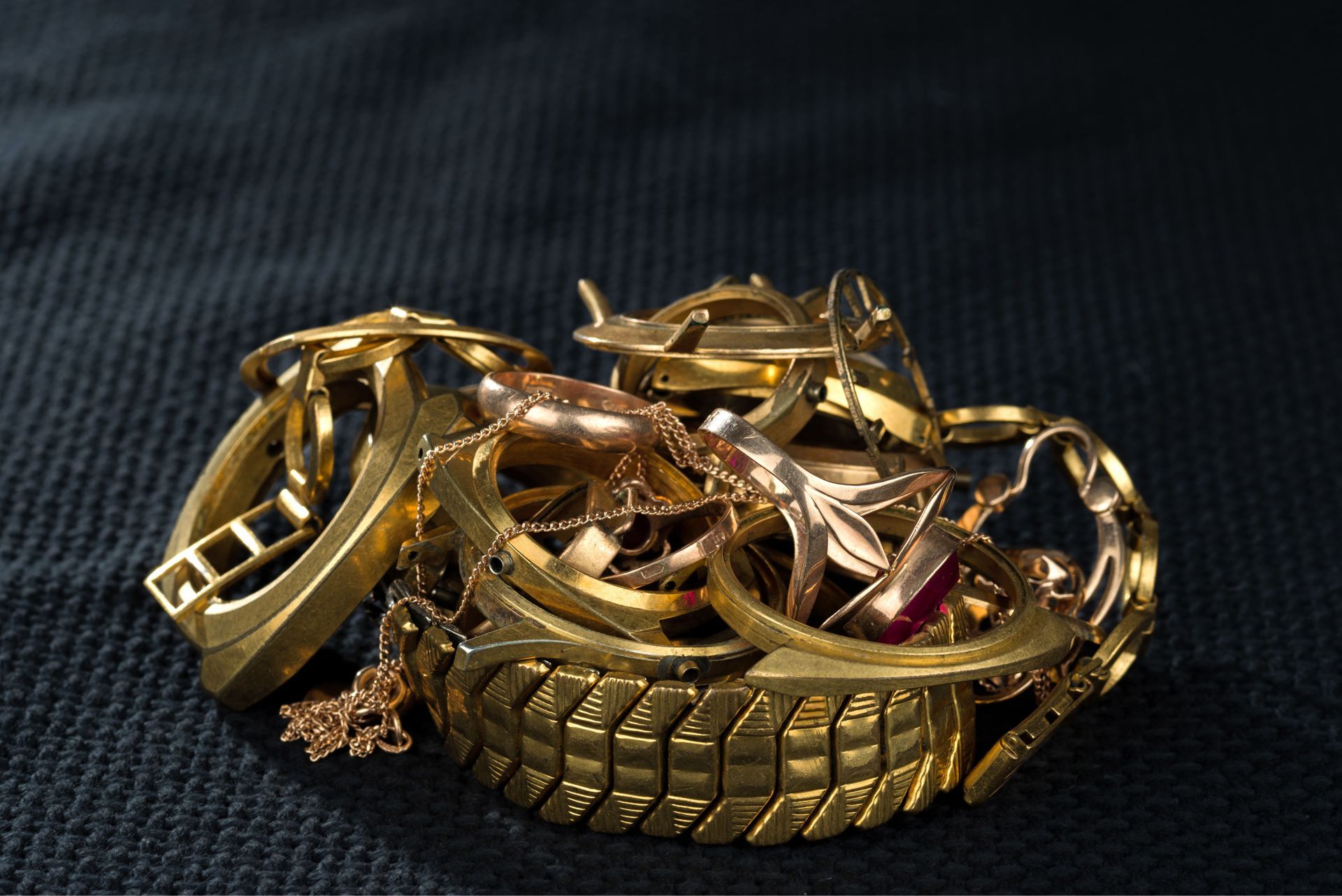 Learn More About Jewelry Recycling A jumbled pile of gold jewelry, including rings, bracelets, and a small charm, set against a dark textured background.