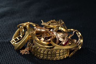 A jumbled pile of gold jewelry, including rings, bracelets, and a small charm, set against a dark textured background.