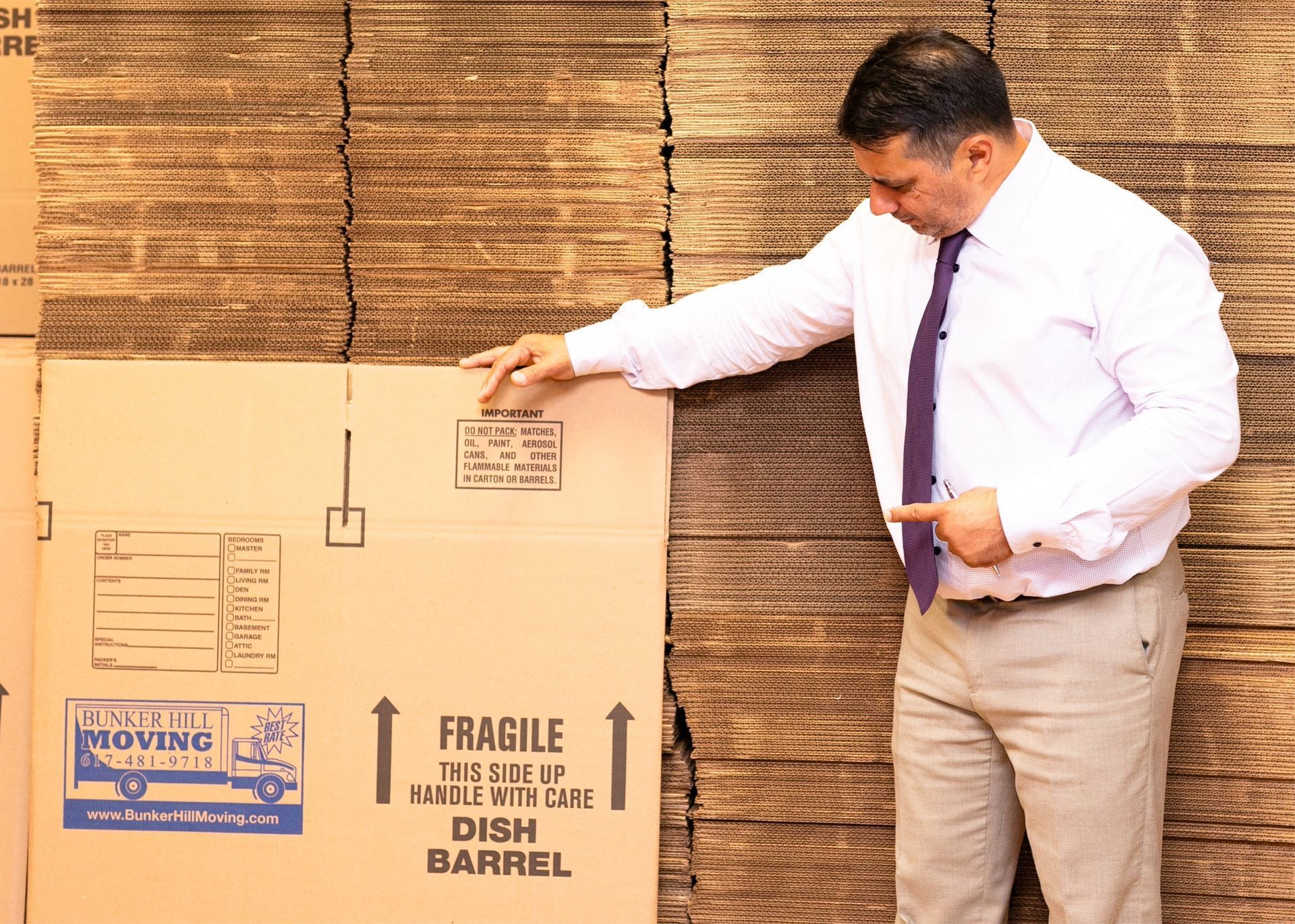 A professional in a white shirt and tie stands in a warehouse, pointing to a cardboard box labeled 