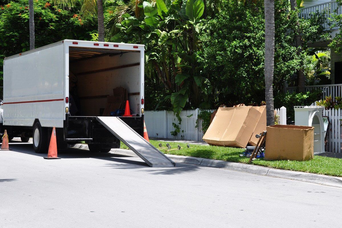 A white moving truck parked on a street with a ramp extended to the curb and cardboard boxes placed on the nearby grass.