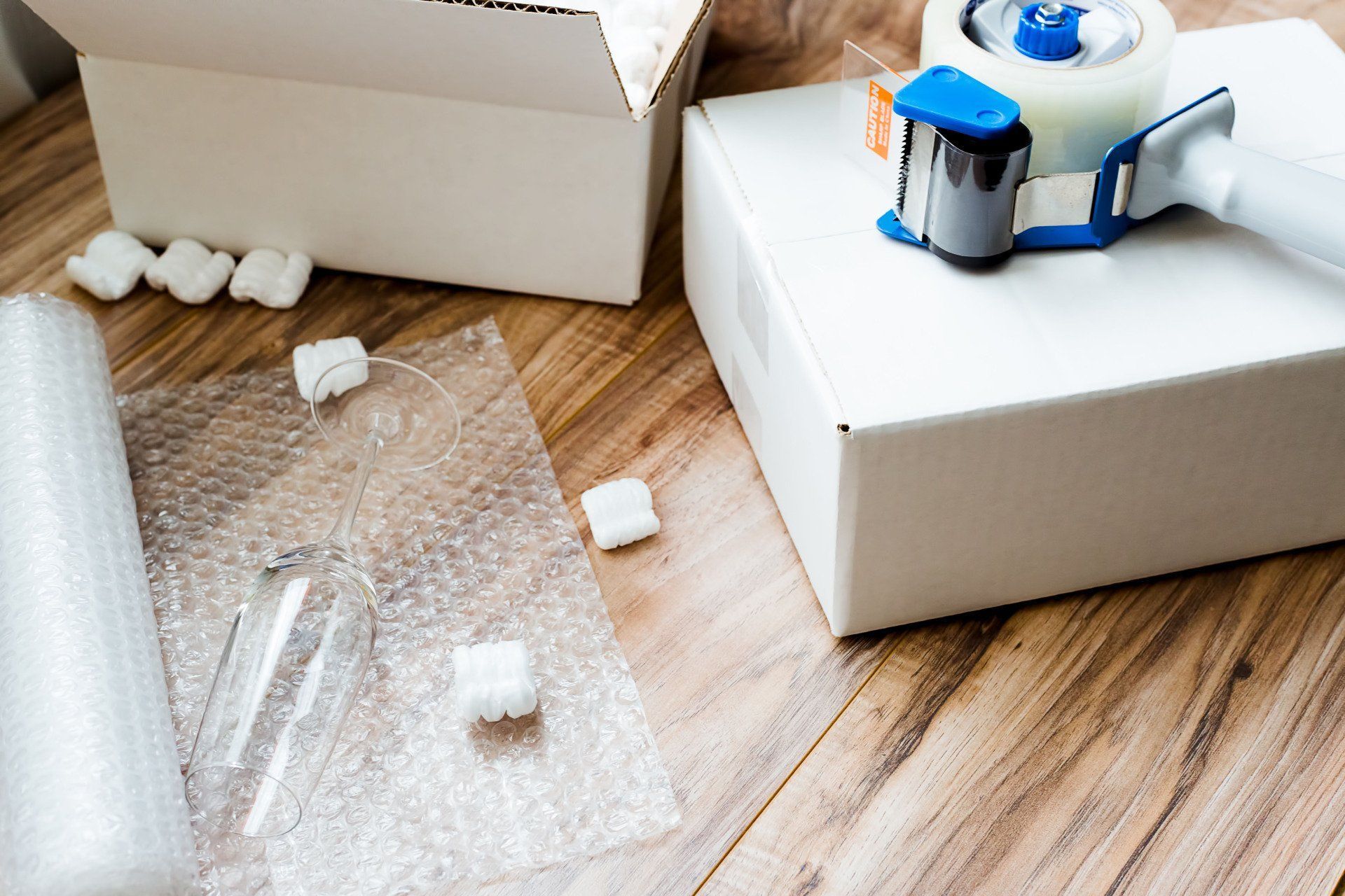 White cardboard boxes, packing peanuts, bubble wrap, and a tape dispenser on a wooden surface with a glass.