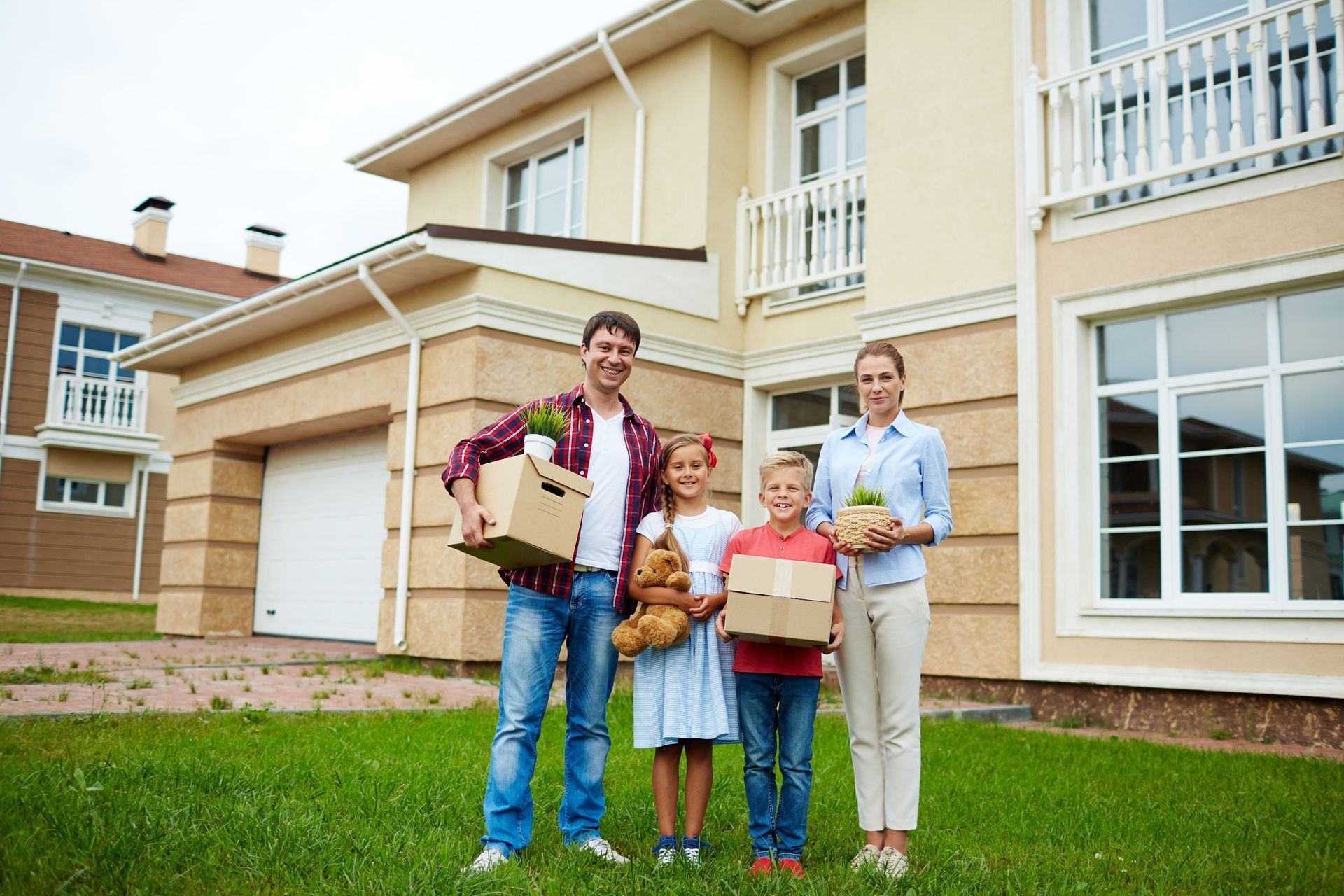 A family stands in front of their new two-story house, holding moving boxes and plants.
