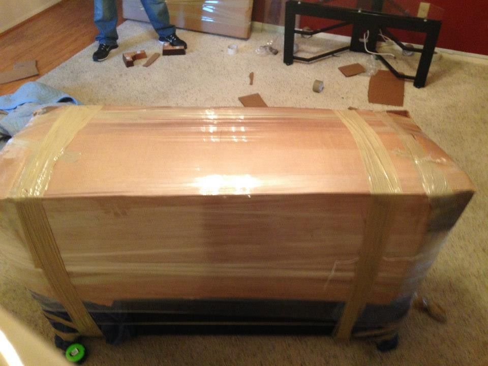 A large, wooden-topped rectangular crate wrapped in clear plastic and packing tape sits on a carpeted floor.