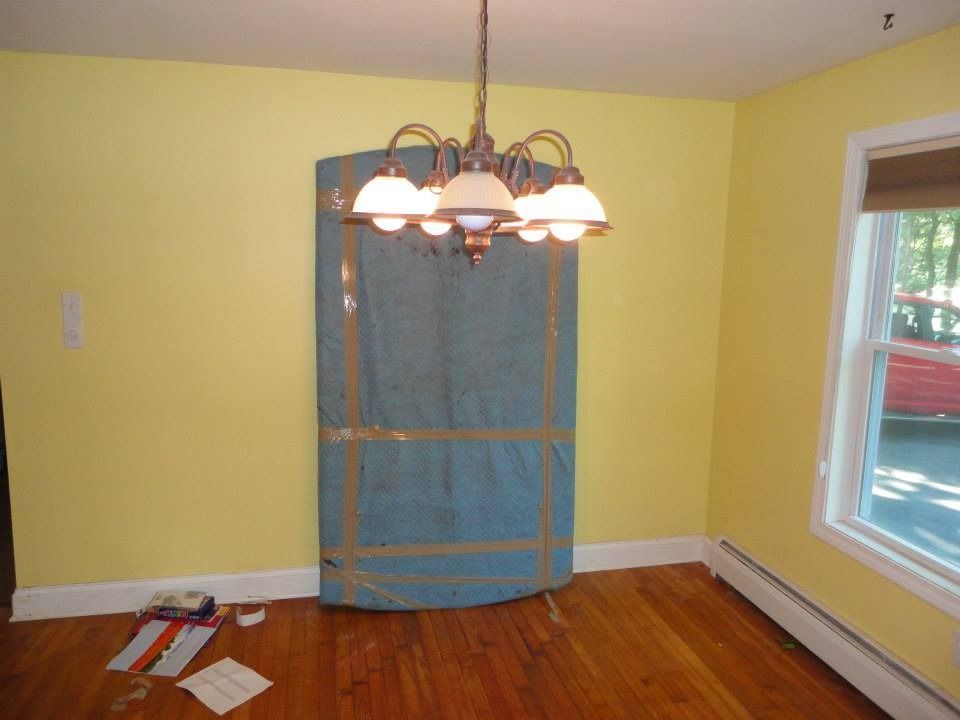 A light yellow dining room with wood floors, a chandelier, and a blue moving-blanket-wrapped piece of furniture.