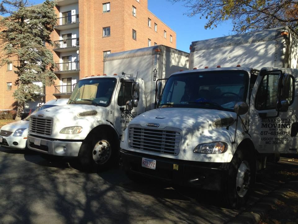 Two white moving trucks parked in a lot next to a brick apartment building on a sunny day.