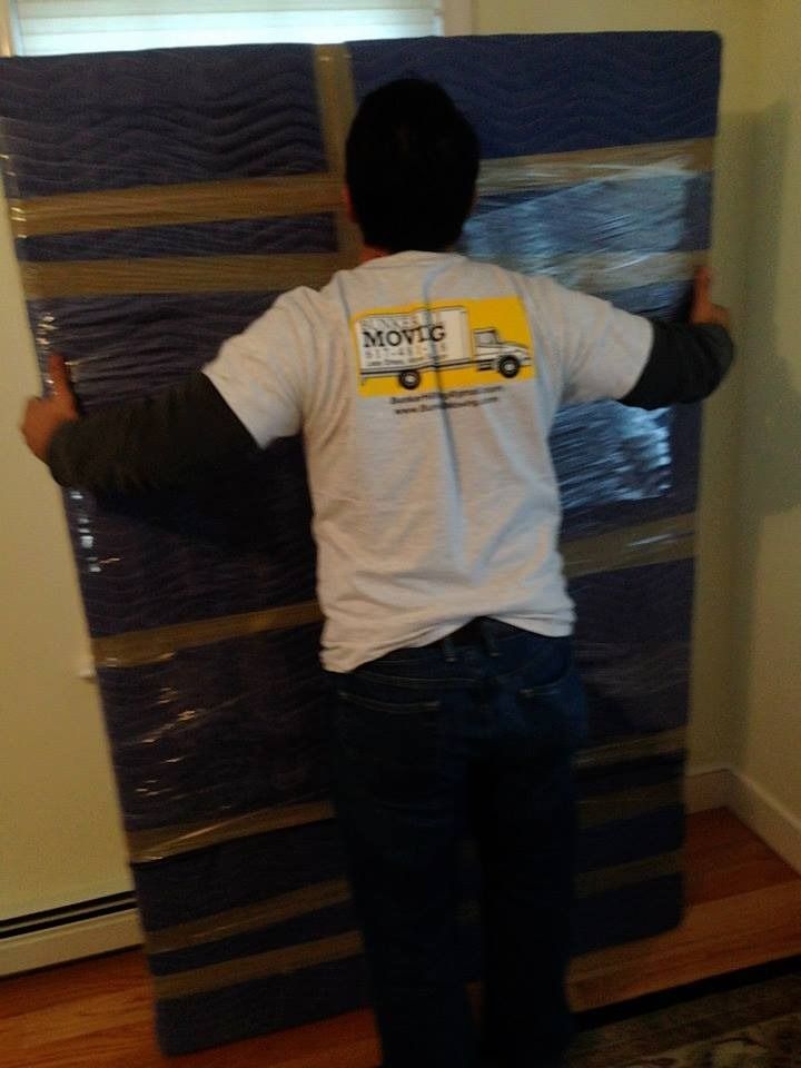 A mover in a gray company shirt carries a large, wrapped item secured with plastic wrap in a room with wood floors.