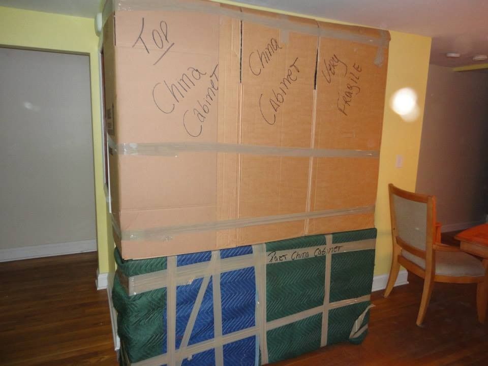 A china cabinet wrapped in cardboard and moving blankets, sitting on a hardwood floor in a room with yellow walls.
