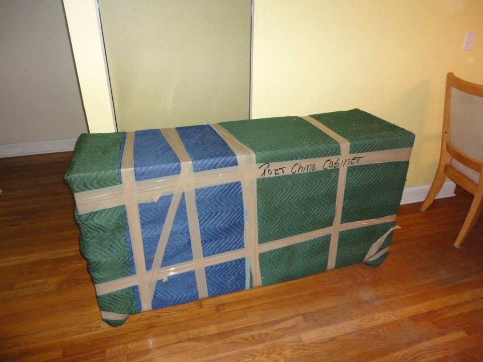 A rectangular piece of furniture wrapped in green and blue moving blankets, secured with packing tape on a wood floor.