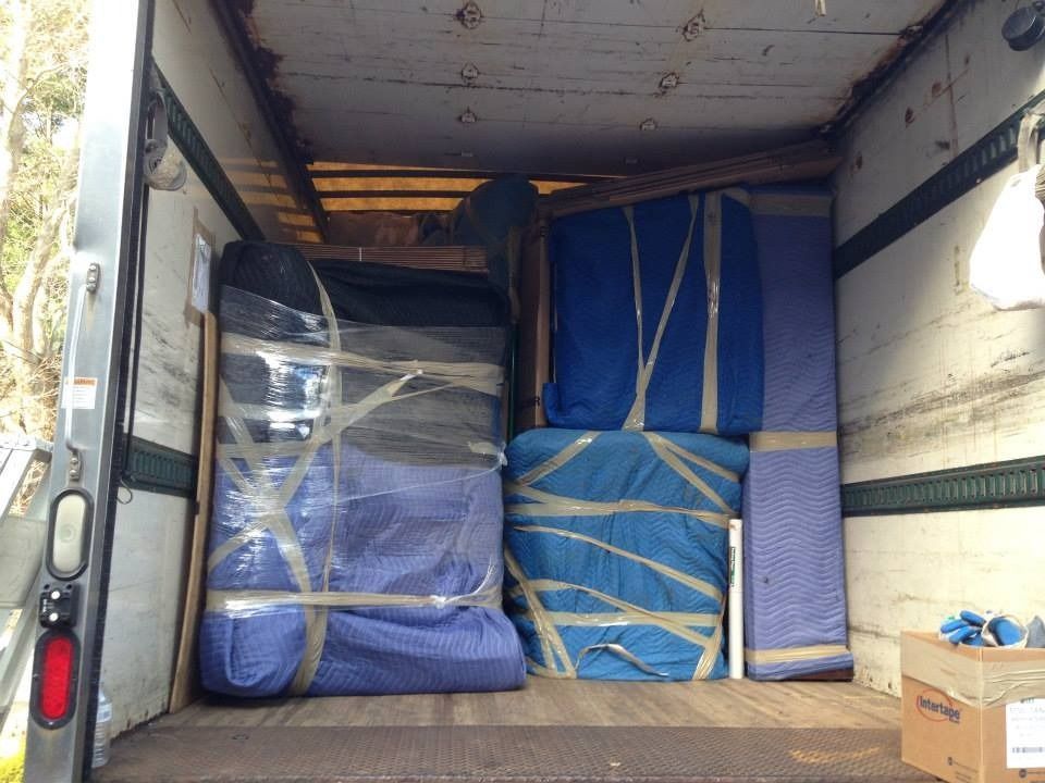 Inside a moving truck, large furniture items are wrapped in blue moving blankets and secured with packing tape.