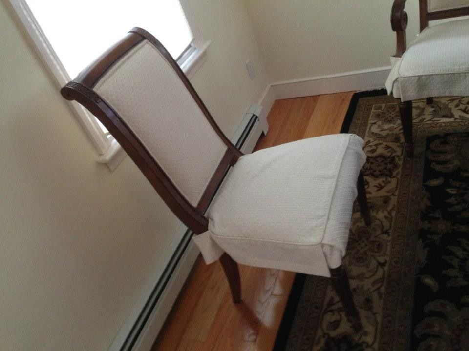 Two dining chairs with dark wooden frames and cream-colored slipcovers standing on a wooden floor near a window.