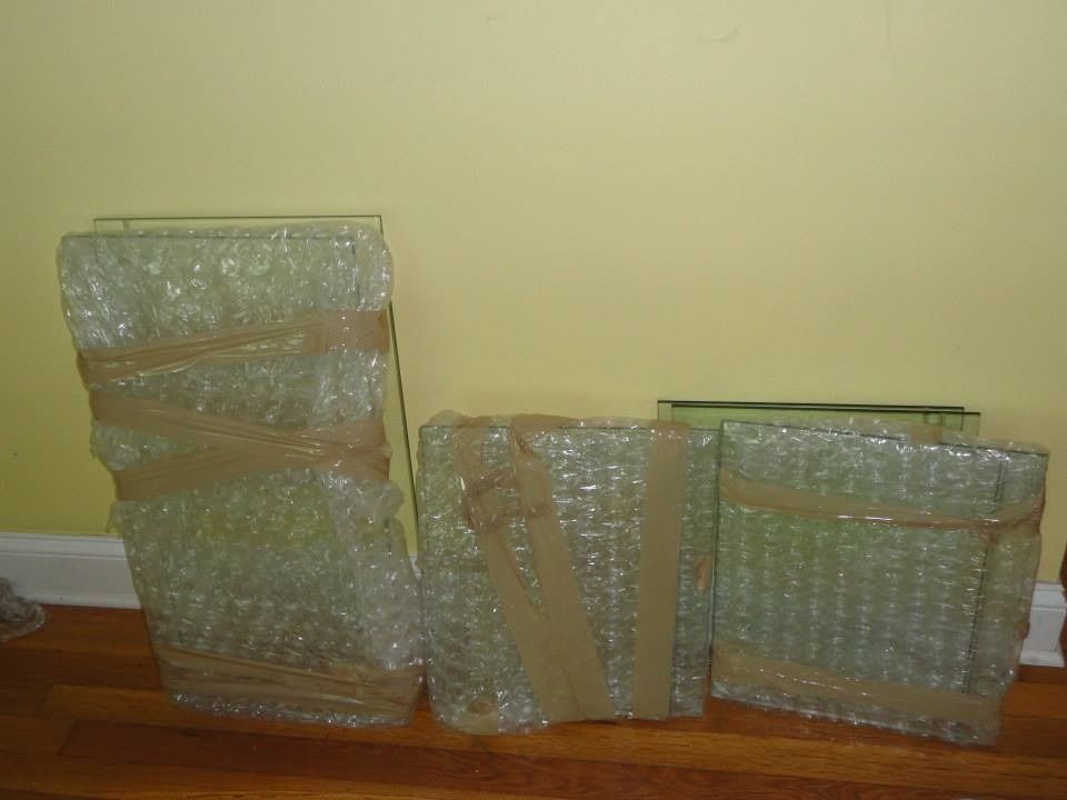 Three glass shelves, wrapped in bubble wrap and secured with tape, stand upright on a wooden floor against a yellow wall.