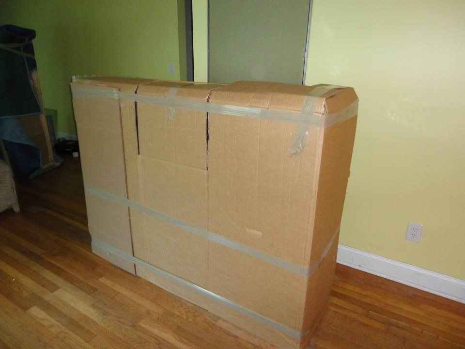 A large rectangular object wrapped in brown cardboard and secured with packing tape, standing on a wooden floor.