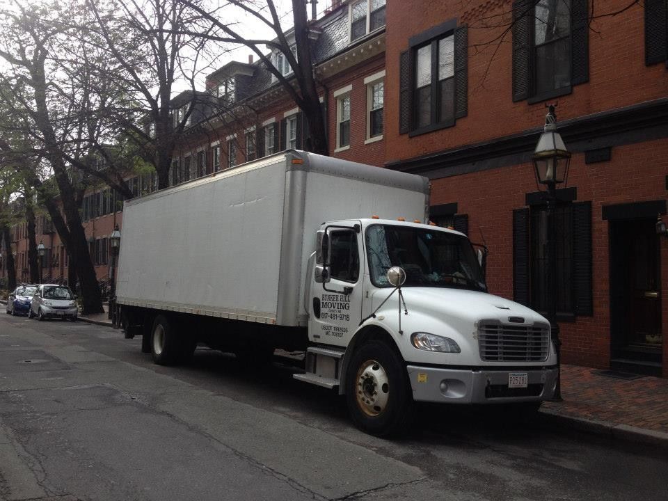A white box truck parked on a narrow city street lined with red brick townhouses and bare trees.