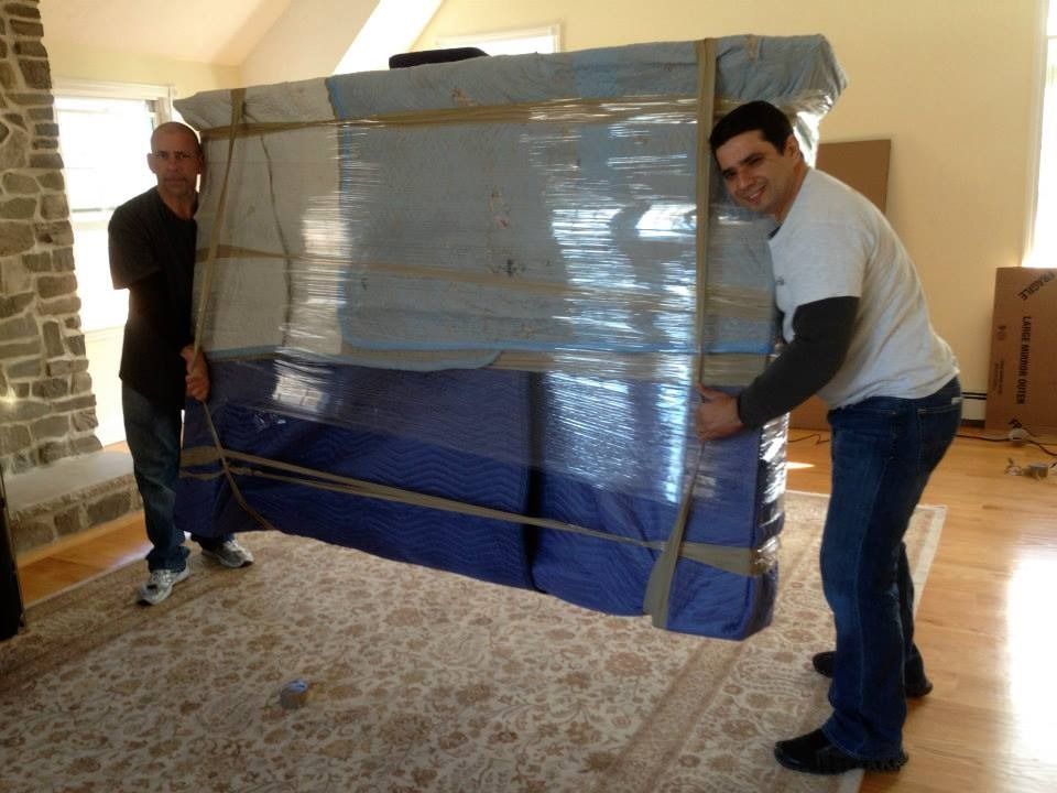 Two people lift a large, plastic-wrapped, blue furniture piece in a brightly lit room with a stone fireplace.