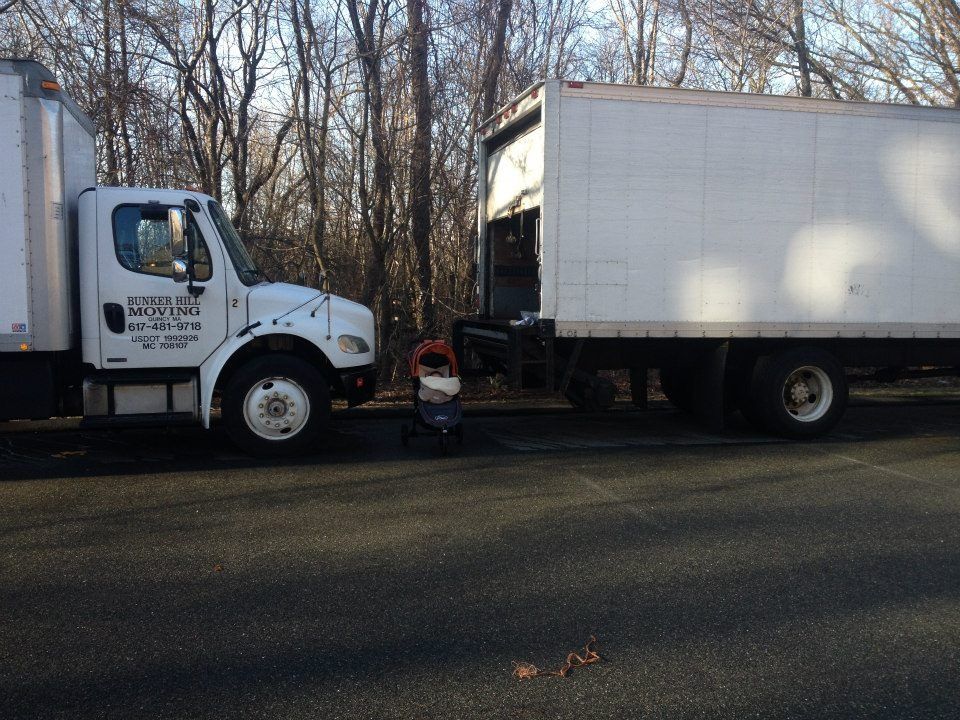 A white box truck parked on asphalt in front of a wooded area, with a baby stroller placed near the side of the cab.