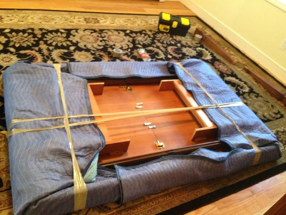 A rectangular table, wrapped in a blue moving blanket and secured with tan packing tape, rests on a patterned rug.