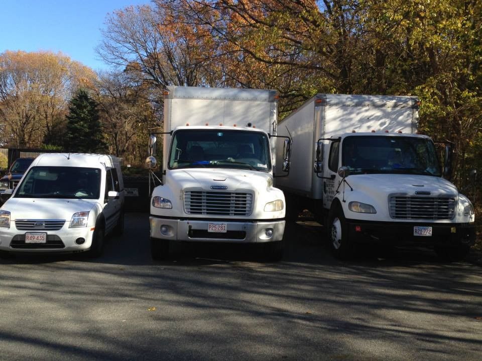Three white vehicles, including two box trucks and a van, parked in a row outdoors against a backdrop of autumn trees.