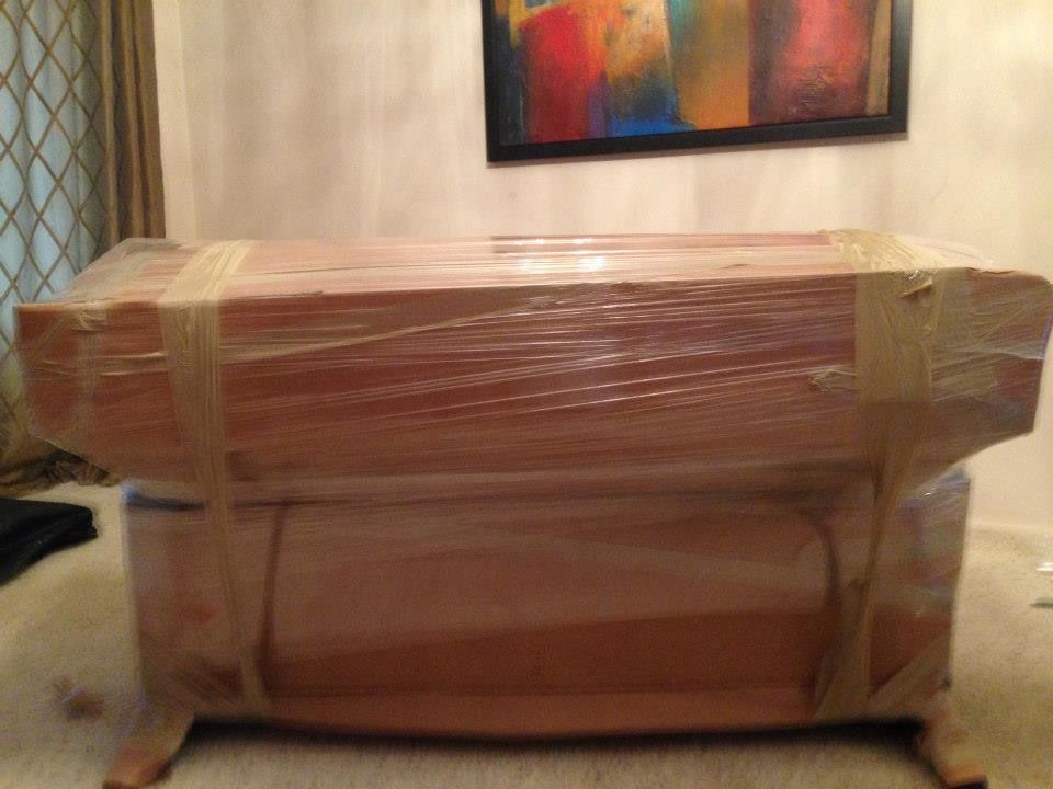 A sofa wrapped in clear plastic and secured with tape, sitting on a carpeted floor in front of a framed painting.