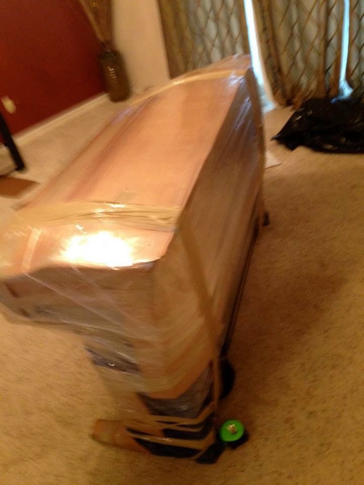 A tall, wood-toned object wrapped tightly in clear plastic film and brown packing tape stands on a carpeted floor.