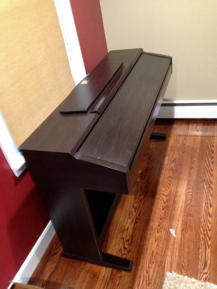 A high-angle view of a dark wood digital piano with a closed lid, standing on hardwood floors in a corner of a room.