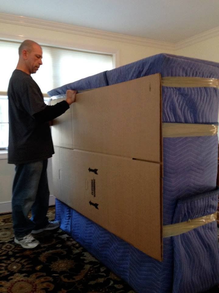 A person securing a cardboard protective panel to the side of a mattress covered in a blue moving blanket.