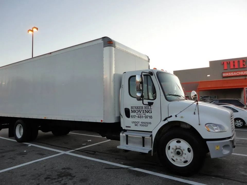 A white moving truck parked in a lot in front of a store building at dusk.