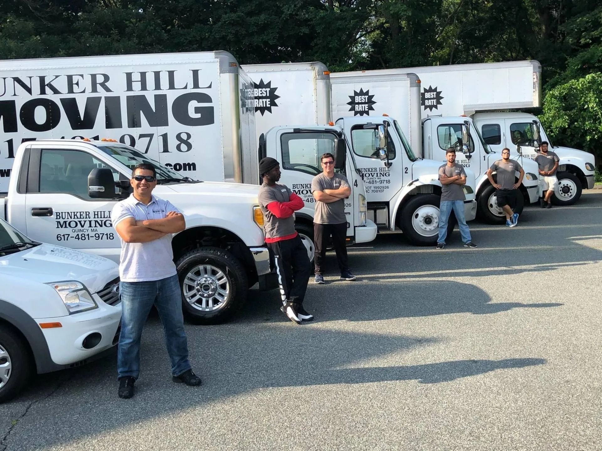 Six people stand with arms crossed in front of a row of Bunker Hill Moving trucks parked in a gravel lot.
