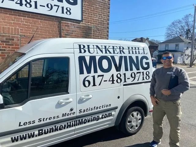 A person stands beside a white Bunker Hill Moving van parked in front of a brick building on a sunny day.