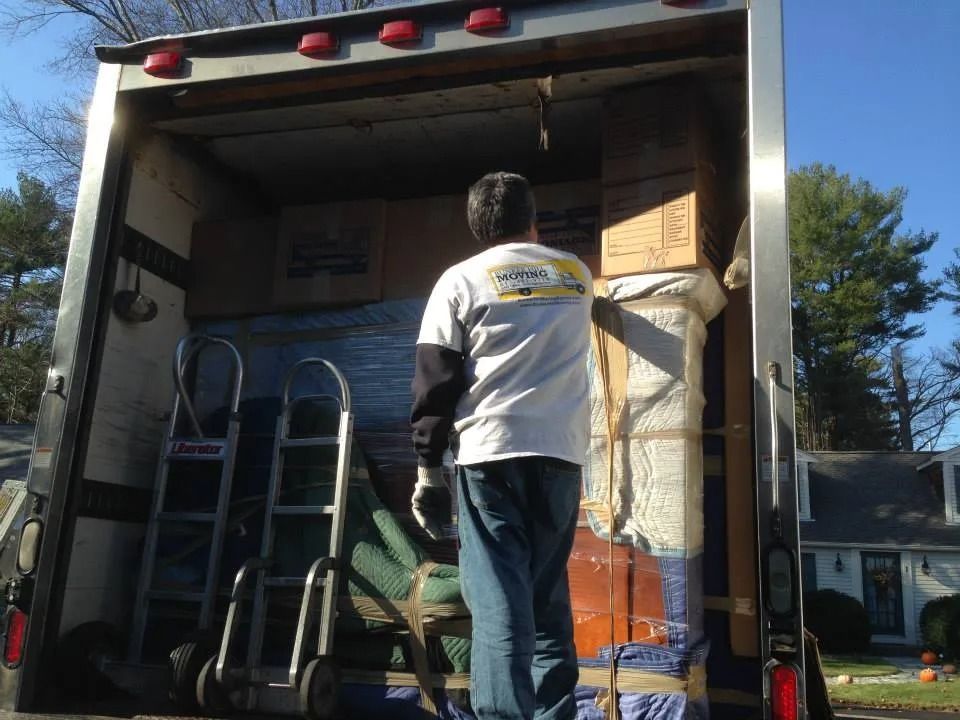 A person wearing a white t-shirt and jeans stands inside the back of a moving truck, loading cardboard boxes and furniture.