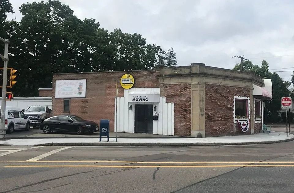 A brick building on a street corner with a white entrance, a yellow circular sign, and parked cars under a cloudy sky.