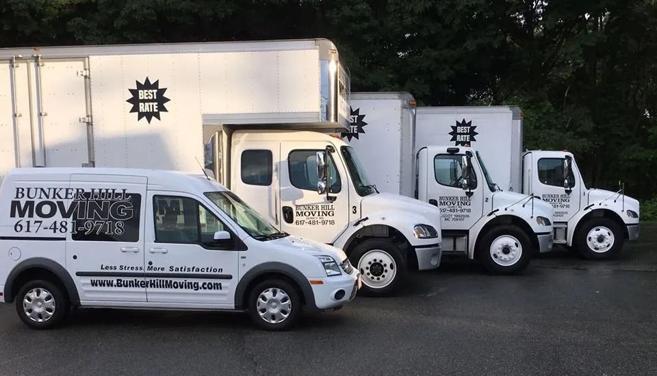 Four white Bunker Hill Moving trucks parked in a row on an asphalt lot against a background of trees.