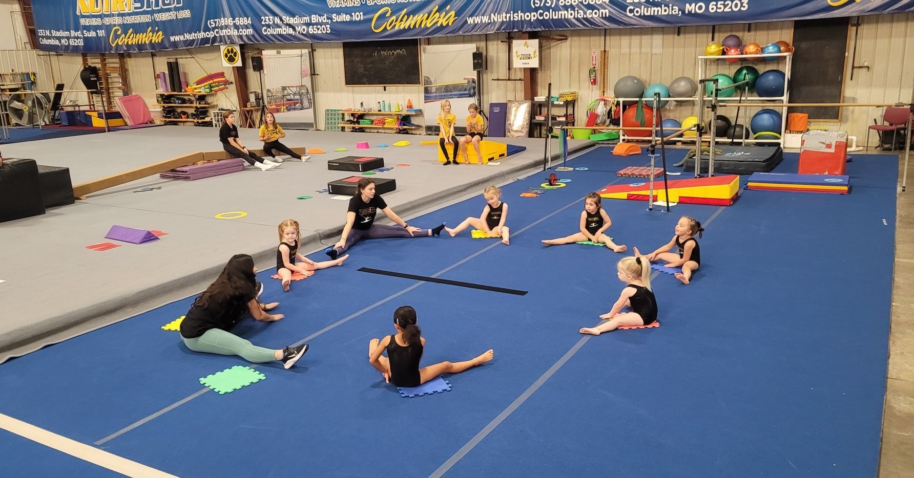 A group of young girls are doing stretching exercises on a blue mat in a gym.