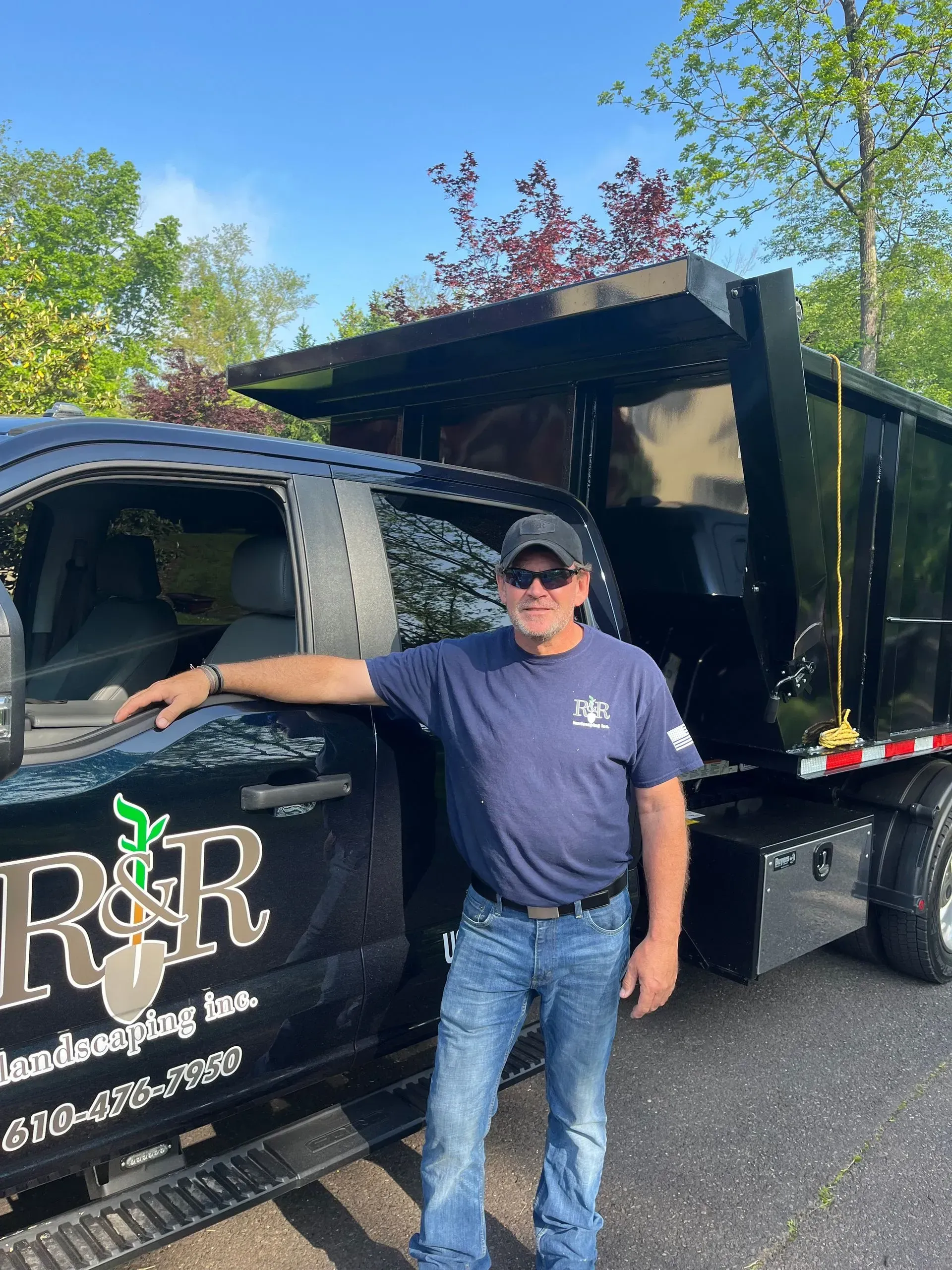Man standing by a black R&R truck and dump trailer in a parking lot, trees in the background