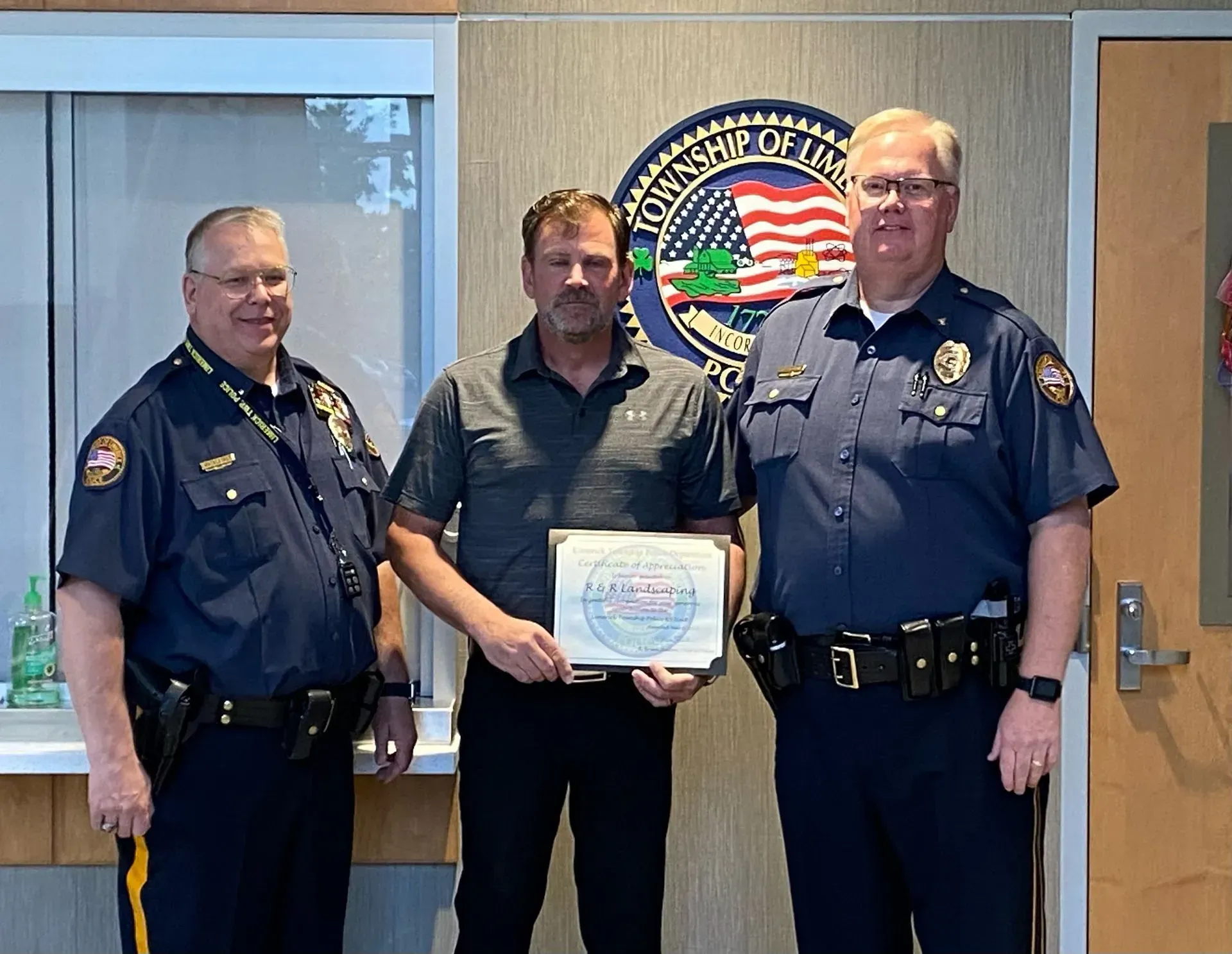 Three men in a police office, with the center man holding a certificate.