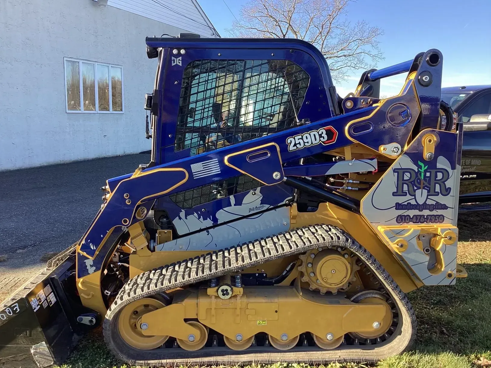 Blue and yellow compact tracked loader parked beside a white building under a clear sky