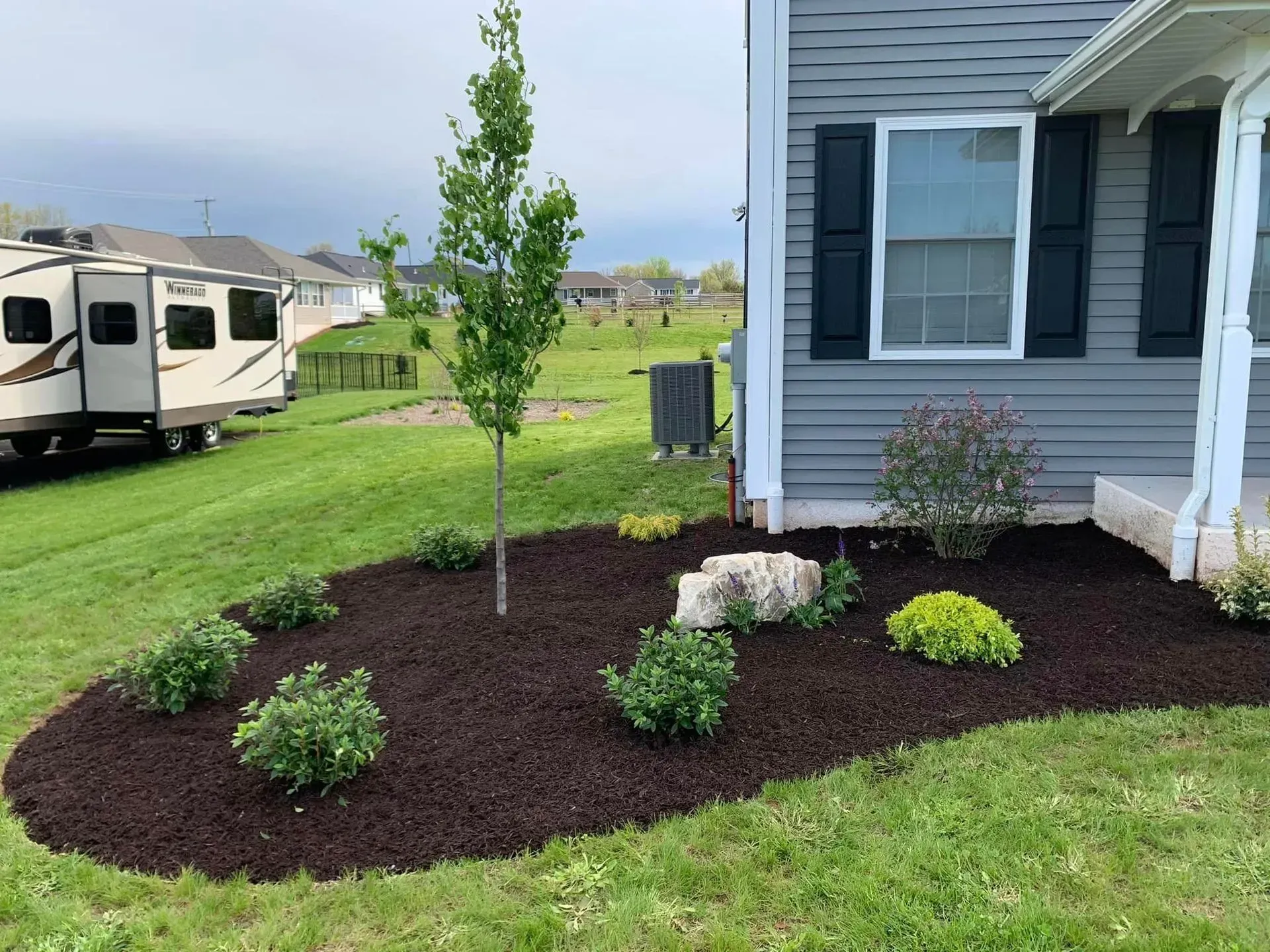Landscaped front yard with new shrubs, mulch beds, and a young tree beside a gray house and parked RV.