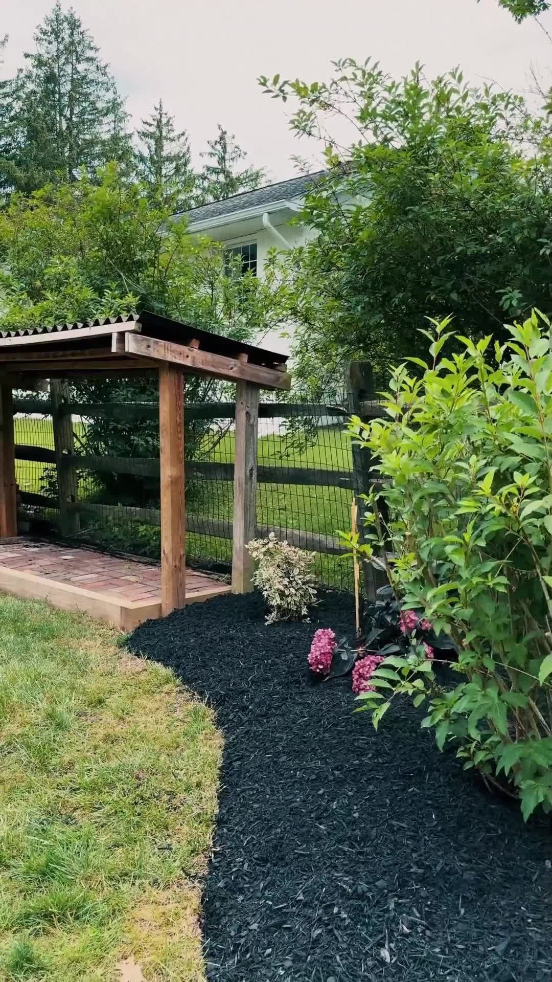 Garden path beside a wooden pergola, bordered by green shrubs and pink flowers, with a house in the background