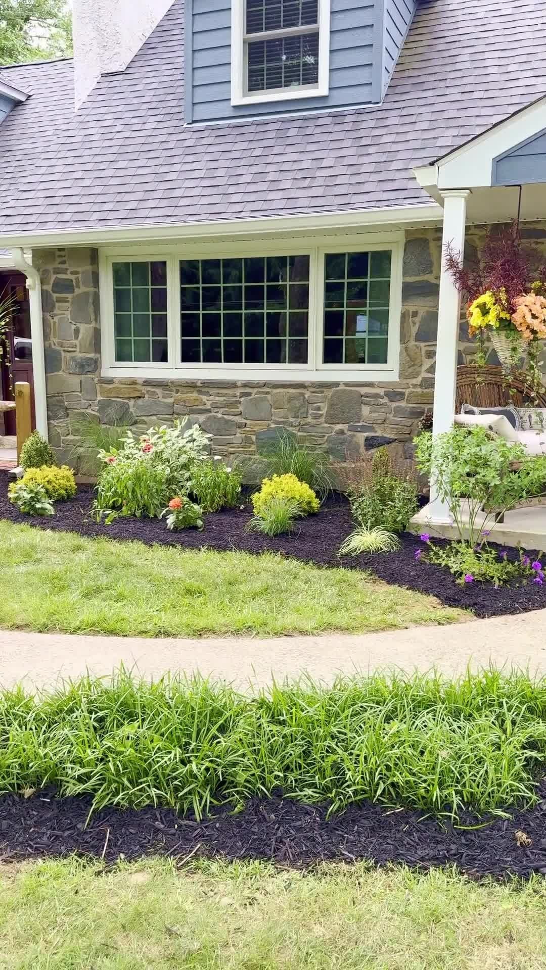 Front yard garden with flowers, shrubs, and a stone house exterior with windows and porch column