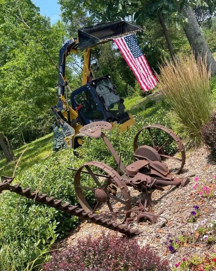 Yellow excavator beside rusted farm implements and an American flag in a garden