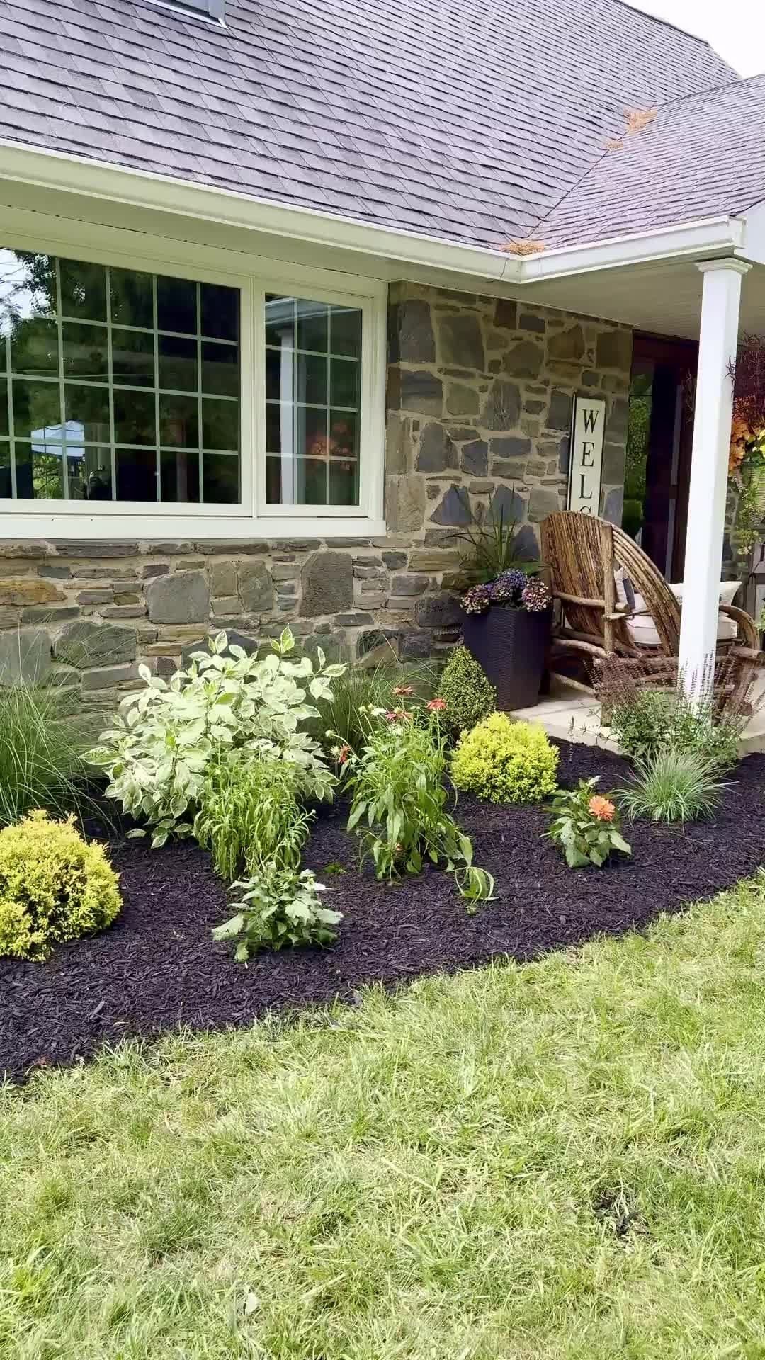 Stone house front with window, porch chair, and fresh landscaped flower bed along the lawn.