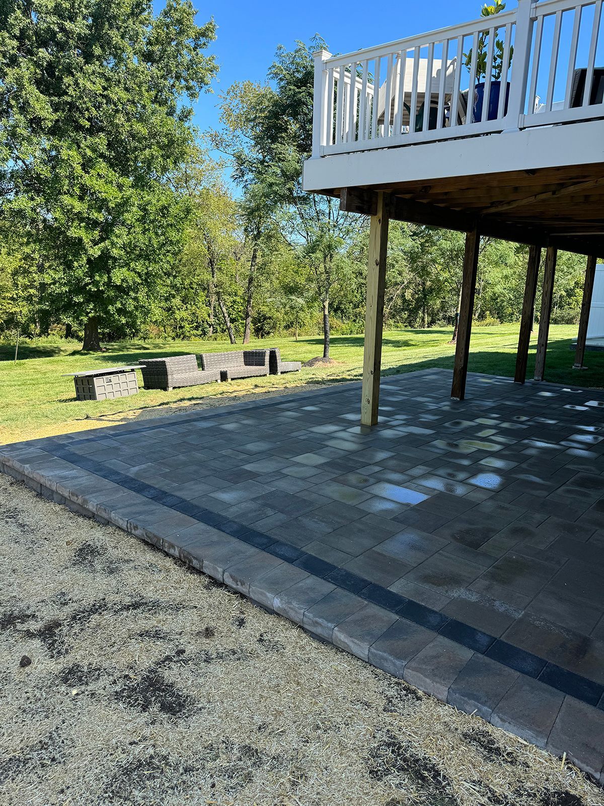Backyard patio under a deck with stone pavers, gravel, and a grassy yard with trees and benches.