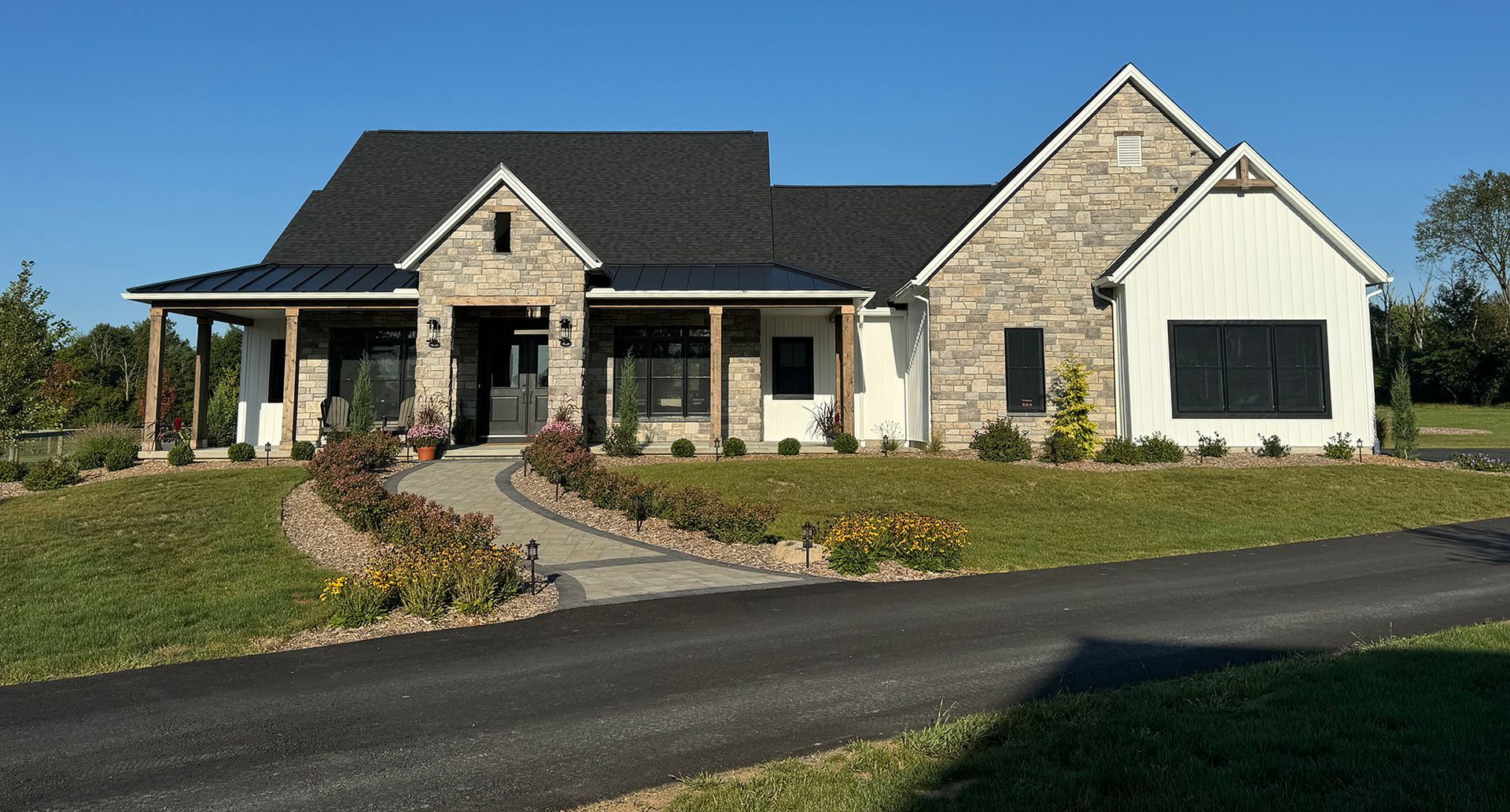 Single-story stone house with a dark roof, front porch, landscaped yard, and curved driveway under a clear sky