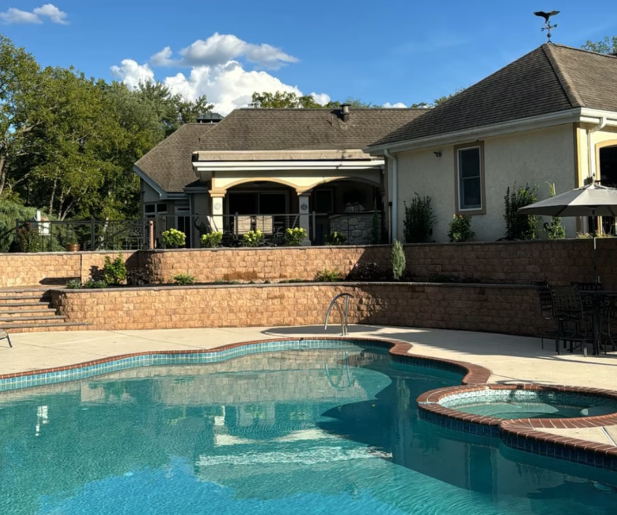 Backyard swimming pool with a brick patio and beige house under a sunny sky
