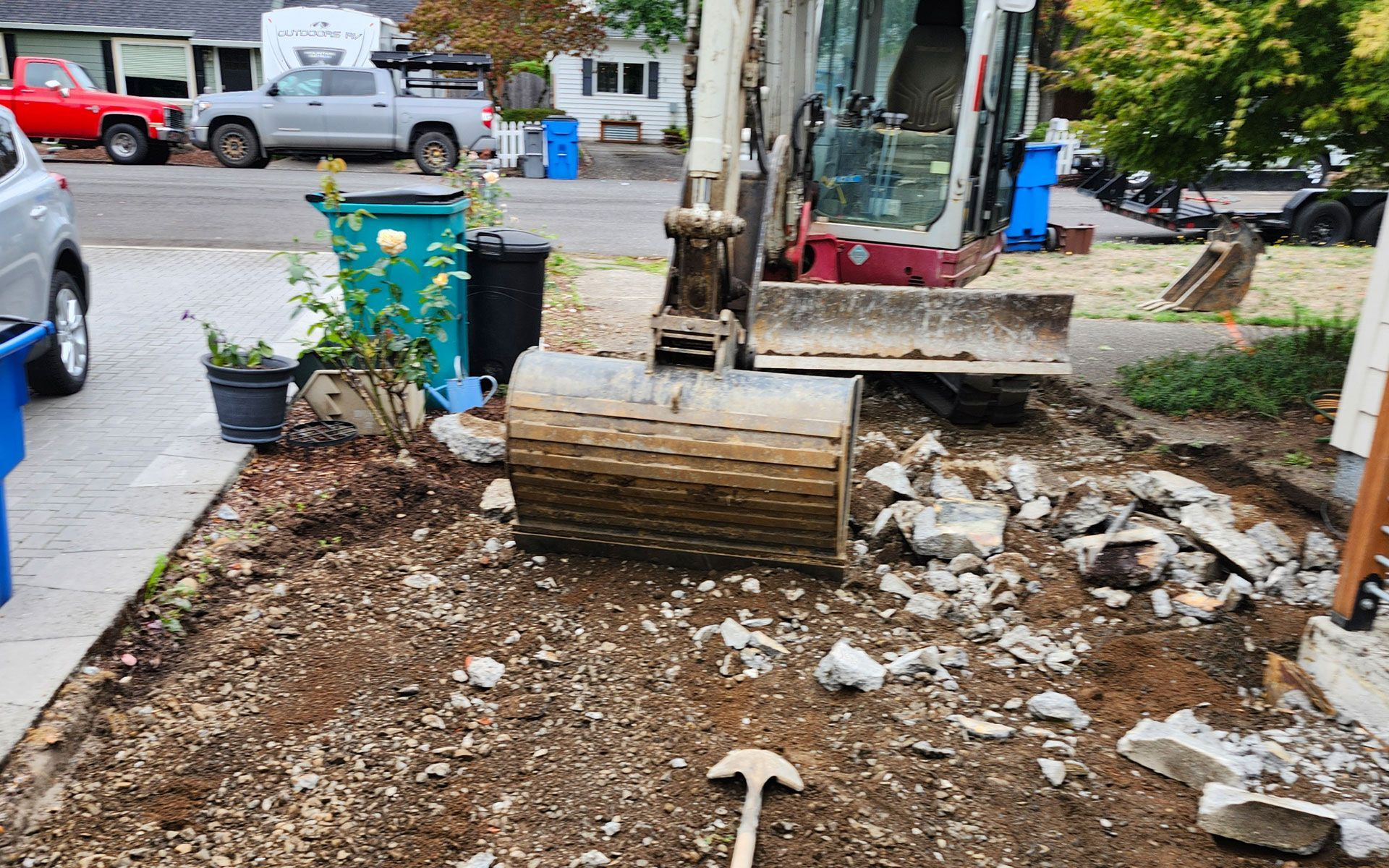Excavator digging up concrete in a residential front yard. Brown dirt, grey concrete debris.