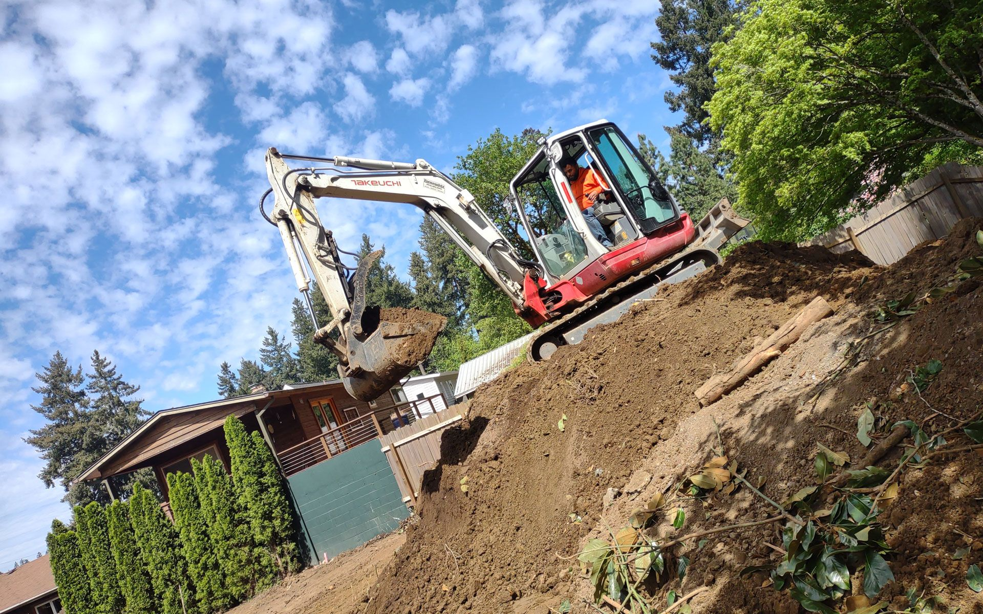 Excavator on a hillside, digging earth with operator visible. Sunny day, blue sky.